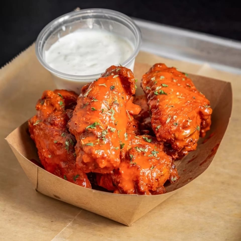 Spicy red chicken wings with herbs in a paper tray, next to a cup of ranch dressing.