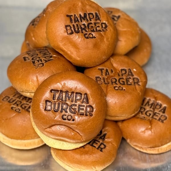 A pile of golden-brown burger buns, each branded with "Tampa Burger Co." on a metallic surface.